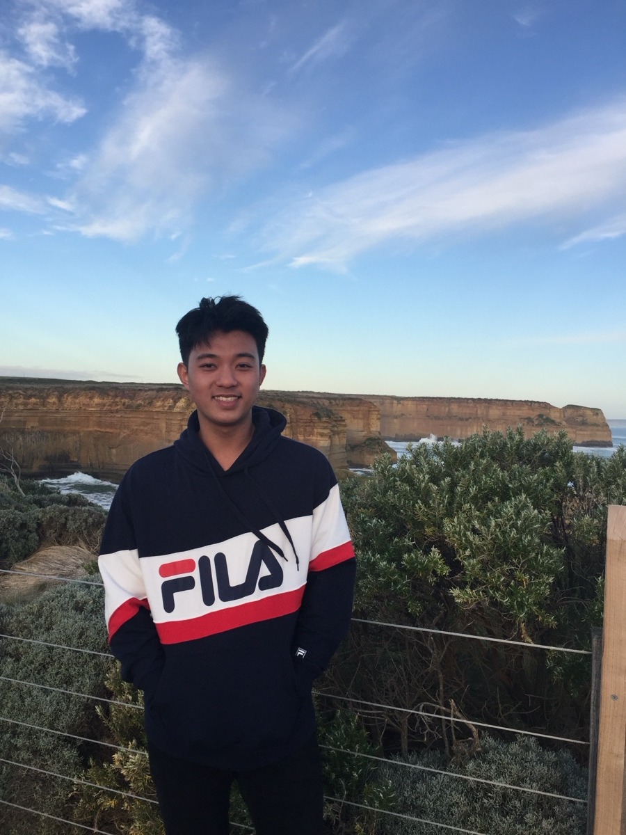 Jarran Tay smiling at a clifftop lookout above the sea, in a navy, white and red hoodie.
